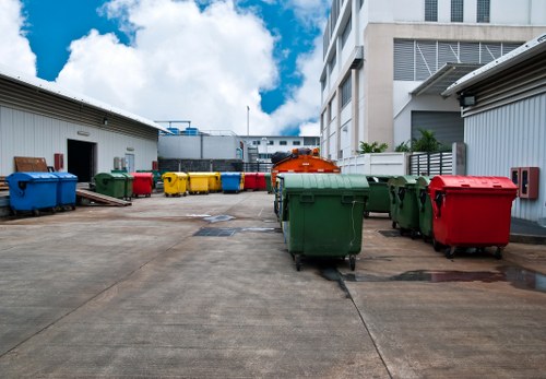 The image shows a paved outdoor area, possibly a service yard, lined with several large wheelie bins in various colours including blue, green, yellow, and red, which are used for rubbish collection. The bins are placed in front of metal storage units or small warehouse structures on either side, with one on the left and another on the right. In the background, there are taller, modern buildings with multiple floors, characteristic of urban commercial or residential complexes, with some windows visible. The sky above is partly cloudy with patches of blue, indicating daytime, and the sunlight illuminates the scene evenly. The setting appears clean and organized, typical of a site managed by a rubbish removal service such as Mill Hill House Clearance, serving the local area near London postcode NW7. The image reflects a typical disposal area where waste is collected before being transported for recycling or disposal, emphasizing the environment of a professional waste management operation.