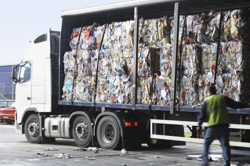 A large commercial refuse truck parked on a paved area, filled with compacted mixed waste paper and cardboard bundles secured within a metal frame. The waste is visibly crumpled, with various colors and textures, including printed and plain materials, creating a chaotic, densely packed appearance. The truck is white with black wheels and rests next to a metallic barrier or railing. A person wearing a high-visibility vest and dark clothing is walking past the vehicle on the right side, with some loose debris scattered on the ground nearby. The background features a clear, bright sky, indicating daylight, and suggests an outdoor setting possibly near a commercial or industrial waste collection site in the vicinity of Mill Hill or the surrounding London boroughs. This image exemplifies the typical process of rubbish collection and transport handled by Mill Hill House Clearance for efficient waste disposal in the area.